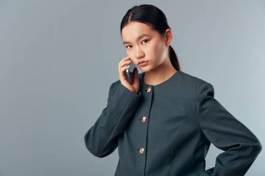 Kendine güvenen, kendine güvenen, koyu renk ceketli, akıllı telefon kullanan genç bir kadın. İletişim ve iş konsepti için el üstünde gri zemine karşı poz veren kadın.