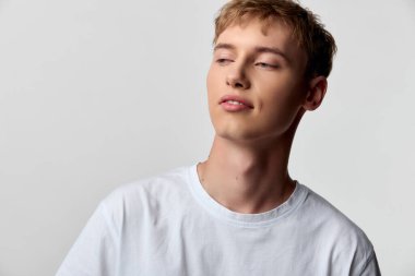 Rahat bir ifade, beyaz tişört, başka tarafa bakan, stüdyo çekimi, çağdaş tarz, gençlik cazibesi, hafif ışıklandırma olan bir erkek modelin yer aldığı sıradan bir moda sahnesi.