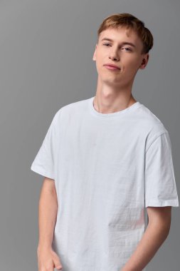 Beyaz tişörtlü genç adam, ışıklandırma, sıradan görünüm ve sakin ifade ile, pazarlama, katalog veya gardırop temalı projeler için uygun minimum bir stüdyo ortamında poz veriyor.