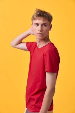 Kırmızı tişörtlü genç adam parlak sarı arka planda duruyor. Düşünce ve merak uyandıran sıradan bir poz. Renk kontrastı ve minimal stil içeren bir stüdyo çekimi.