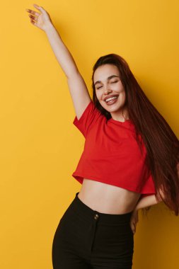 Uzun kahverengi saçlı, kırmızı tişörtlü ve siyah pantolonlu mutlu bir kadın. Kapalı gözlerle gülümsüyor. Pozitif ruh hali için açık sarı arka planda koluyla poz veriyor..