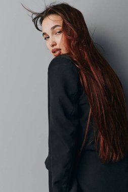 Gri arka planda duran düz uzun saçlı ve siyah giysili genç bir kadın. Makyajı az olan, kendine güvenen ve rahat bir şekilde kameraya bakan bir kadın model..