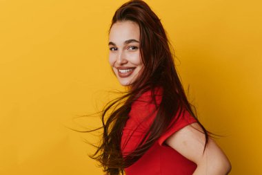 Koyu uzun saçlı, kırmızı bluzlu, parlak sarı arka planda omzunun üstünden bakan gülümseyen bir kadın. Neşeli yüzü ve günlük tarzı olan, yakın çekim stüdyo portresi olan bir kadın model..