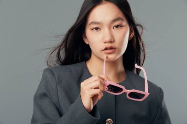 Gri ceketli, düz saçlı, ağzı yakınında pembe gözlüklü genç bir kadın güzellik portresi için yumuşak ışıklandırmayla tarafsız arkaplanda kameraya bakıyor..