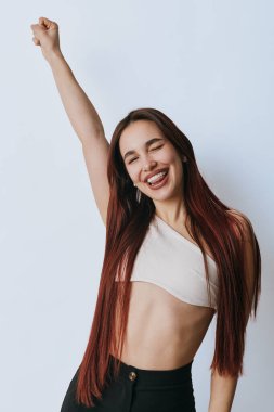 Uzun saçlı, beyaz saçlı, siyah pantolonlu gülümseyen genç bir kadın sevinçle kolunu kaldırıyor, özgüvenini ve pozitif duygularını arka planda gösteriyor..