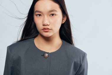 Gri resmi ceket giyen, düz siyah saçlı genç bir kadın stüdyoda ciddi ve sakin görünüyor. Tarz ve profesyonel görünüş için sade arkaplanı olan güzel bir portre.