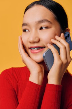 Telli, siyah saçlı, kulağının yanında cep telefonu tutan gülümseyen bir kadın. Elleri yanağında kırmızı örgü giysiler giyiyor. Hayat tarzı ve iletişim için sağlam sarı bir zemin üzerinde yakın plan resmi var.