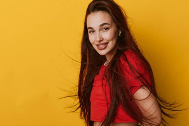 Gülümseyen genç bir kadın, dalgalı saçlarıyla kırmızı bluzuyla canlı sarı arka planda poz veriyor. Kendine güveni tam bir kadın portresi ve parlak bir ortamda rahat bir duruş..