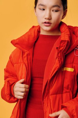 Kırmızı kabarık ceketli ve uyumlu elbiseli, turuncu arka planda duran genç bir kadının portresi. Şık kış kıyafetleri, kadınlar için sıcak ve renkli mevsimlik kıyafetlere yakın çekim..