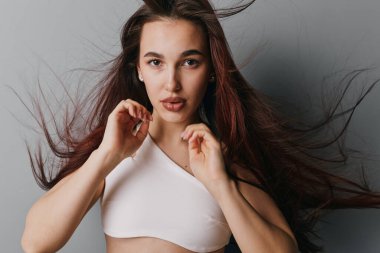 Saçları dökülmüş, beyaz bluzu olan ve kameraya bakan genç bir kadının portresi. Stüdyo ortamındaki gri arka planda doğal görünümlü ve yumuşak ışıklandırmalı kadın güzellik modeli..