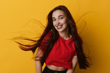 Kırmızı bluzlu gülümseyen genç kadın sarı arka planda poz veriyor. Uzun saçlı, hareketli bir kadın portresi, neşeli bir ifade, ve parlak stüdyo ortamında günlük kıyafetler..