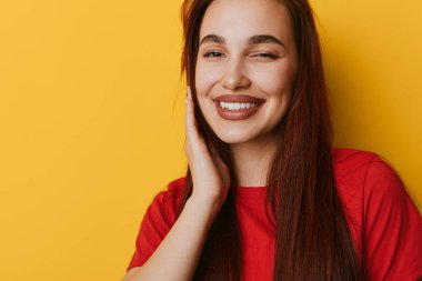 Uzun kahverengi saçlı, gülümseyen kadın açık sarı arka planda kırmızı bluz giyiyor. Elleri yanağında, olumlu duygu ve güzelliği gösteren neşeli bir kadın portresi..