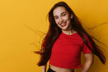 Sarı duvarın önünde kırmızı bluz giyen, uzun saçlı, gülümseyen genç bir kadın. Neşeli kadın portresi. Parlak bir ortamda doğal bir ifade ve günlük kıyafetler..