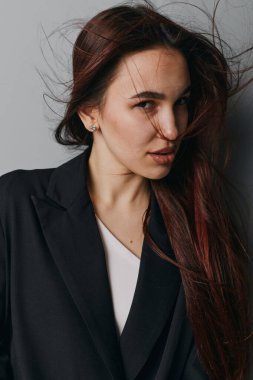 Koyu renk uzun saçlı, odaklanmış siyah ceketli ve beyaz gömlekli genç bir kadın. Minimalist gri arkaplan ve gündelik zarif tarzlı stüdyo portresi.