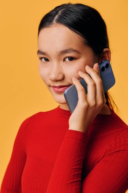 Kırmızı kazaklı, gülümseyen bir kadın cep telefonuyla yüz yüze konuşuyor. Parlak turuncu arkaplandaki yakın plan portresi teknoloji kullanımı, modern iletişim ve dost ifadesini gösteriyor.