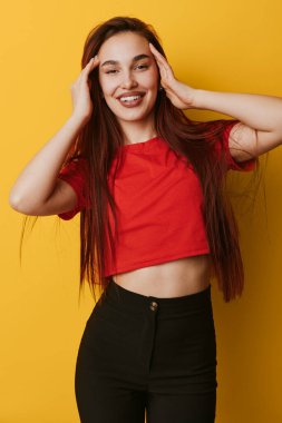 Kırmızı gömlekli, siyah pantolonlu, sarı arka planda duran ve yüzüne dokunan mutlu bir kadın manken. Stüdyoda gençlik tarzı, güven ve neşeli ifade.