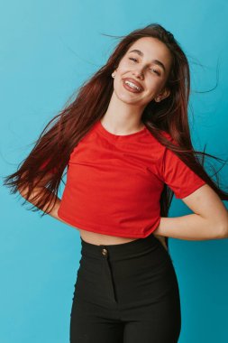 Kırmızı gömlekli, uzun kahverengi saçlı ve siyah pantolonlu gülümseyen kız mavi arka planda güven içinde poz veriyor. Neşeli bir kadın portresi, günlük kıyafetler ve doğal bir gülümseme..