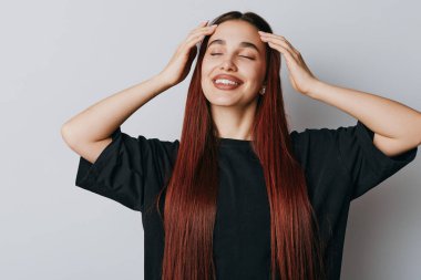 Düz kızıl saçlı, gözleri kapalı gülümseyen ve elleri kafasında siyah bir bluz olan genç bir kadın. Sakin duygusal ifade, pürüzsüz cilt ve basit stüdyo arka planı.