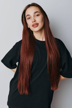 Siyah tişört giyen uzun kızıl saçlı genç bir kadın gri arka planda duruyor. Saç stili, güzellik, portre, günlük kıyafetler ve en az makyajla özgüven ifadesi..