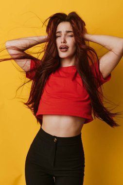 Kırmızı gömlekli, siyah pantolonlu, saçları dalgalı, başı sarı arka planda kapalı genç bir kadın. Etkileyici bir duruş, modern kıyafetler ve yaşam tarzı ve moda için canlı bir renk..
