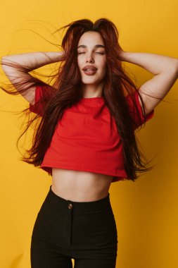Kızıl saçlı, gevşek suratlı, kırmızı gömlekli, siyah pantolonlu, sarı arka planda duran kadın manken. Fotoğrafta yaşam tarzı, gençlik ve güven kavramı.