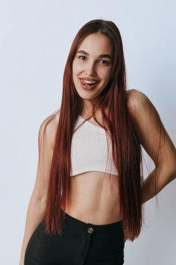Uzun kızıl saçlı, beyaz kolsuz bluz ve siyah pantolon giyen gülümseyen genç bir kadının portresi. Dişi model, rahat ve mutlu bir ruh haliyle, düz ışık duvarına karşı el üstünde poz veriyor..