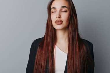 Uzun kızıl saçlı, siyah ceketli ve beyaz bluzlu, gözleri kapalı genç bir kadının portresi. Pürüzsüz ciltli ve doğal görünüşlü bir kadın. Düz gri zemine karşı duruyor..