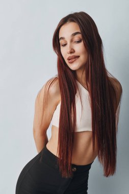 Uzun örgülü, beyaz kolsuz bluzlu ve siyah pantolonlu genç bir kadının portresi. Gündelik tarz, güzellik ve modern saç stili. Stüdyoda yumuşak ışıklandırmayla çekildi..