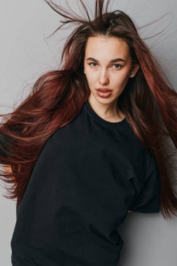 Gri duvarın önünde dalgalı kızıl saçlı ve siyah gömlekli genç bir kadın portresi, kendine güvenen bir görünüm ve moda veya güzellik konsepti için ince bir makyaj sergiliyor..