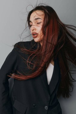Siyah ceketli, kırmızı saçlı, gözleri kapalı, saçları hareketli bir kadın portresi. Tarafsız arkaplanlı bir stüdyo çekimi. Şık, tarz ve güzelliği sıradan moda tarzıyla gösteriyor..