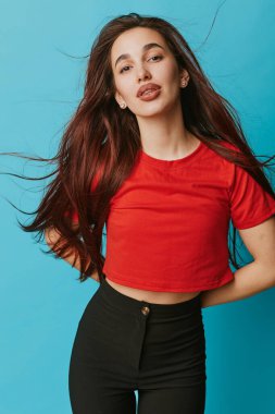 Kırmızı kısa kollu gömlekli kadın model ve mavi arka planda poz veren yüksek bel siyah pantolon. Uzun kahverengi saç, minimal aksesuar, günlük kıyafet, güzellik ve gençlik konsepti stüdyoda..