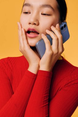Kırmızı balıkçı yaka kazaklı Asyalı kadının yüz yüze telefonla konuşması, duygu ve tepkileri aktarması, açık sarı arka planda izole edilmesi ve metin için metin alanı bırakılması.