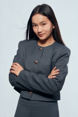 Uzun siyah saçlı, gri takım elbiseli, kollarını kavuşturmuş, resmi kıyafet ve kendine güvenen yüz ifadesi için hafif arka planda duran bir iş kadını..