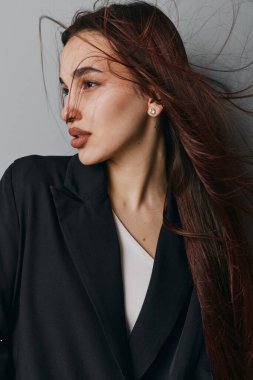 Siyah ceketli, düz saçlı, beyaz bluzlu, yan bakışlı genç bir manken. Minimalist stil, doğal görünüş, küpeler ve saçlar, tarafsız zemine hafifçe esiyor..