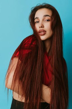 Uzun kahverengi saçlı ve minimum makyajlı kırmızı gömlekli genç bir kadın portresi. Mavi arka planlı stüdyo çekimi, çapraz kollar pozu ve moda veya güzellik için rahat yüz ifadesi..