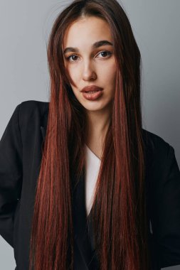 Uzun kahverengi saçlı, siyah ceketli, beyaz gömlekli, kendine güvenen genç bir kadının portresi gri duvarın önünde duruyor..