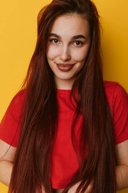 Kırmızı bluzlu, uzun saçlı, sırıtan genç bir kadın sarı arka fonun önünde dikiliyor. Kapalı mekanda doğal ifadesi ve günlük tarzı olan neşeli bir kadın portresi..