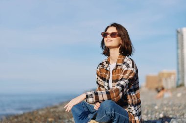 Sahilde güneş gözlüklü, yaz güneşinin, deniz melteminin ve sakin bir sahil havasının tadını çıkaran kendine güvenen bir kadının olduğu sıradan, şık bir açık hava sahnesi..