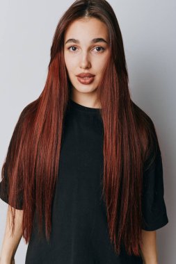Uzun kızıl saçlı ve sıradan siyah tişörtlü genç bir kadın portresi. Moda ve güzellik anlayışı için doğal görünüşü ve sakin yüzü olan düz bir duvarın önünde poz veriyor..