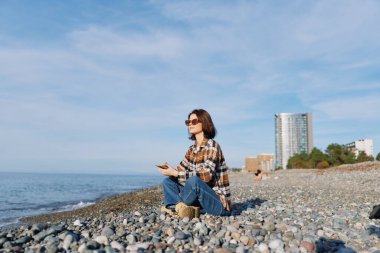 Kayalık bir sahilde, güneş gözlüklü, kot pantolonlu ve ekose bluzlu, sakin bir sahil şeridi boyunca deniz kenarının tadını çıkaran ve mavi ufuk çizgisi olan bir kadın..