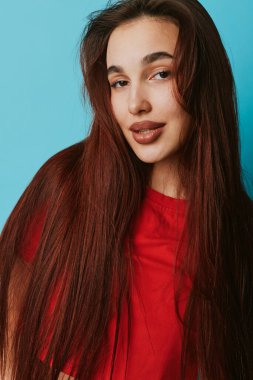 Mavi duvara karşı düz kahverengi saçlı, gülümseyen genç bir kadının portresi, doğal görünüş, gevşek ifade ve güzellik için sağlıklı cilt ve günlük moda..