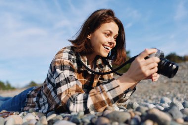 Kayalık bir sahilde bir kadın fotoğrafçının olduğu açık hava sahnesi, elinde kamera, parlak ışık altında gülümseme, günlük kıyafet, hobi fotoğrafçılığı ve doğanın samimi anı, yaratıcı boş vakitleri aktarma ve
