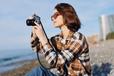 Sahilde bir kadın fotoğrafçının açık hava portresi, elinde kamera, güneşli sahil şeridi, ekose üstlü, sakin okyanus, hafif esinti, kompozisyon, teknik ve doğa boyunca keşfine odaklan.