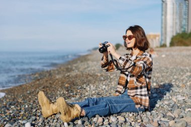 Kayalık bir sahilde kameralı sıradan bir gezgin. Dalgalar ve uzak binalar, açık havanın, parlak güneşin, temiz havanın ve deniz kenarında yaratıcı bir anın tadını çıkarıyor. Yaşam tarzını ve doğayı yakalıyor.