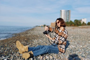 Açık deniz fotoğrafçısı portresi, ufku hedefleyen lensler, güneşli deniz ve kayalık kıyı, günlük kıyafetler, yaratıcı fotoğrafçılık, seyahat titreşimleri ve deniz kenarı keşifleri.