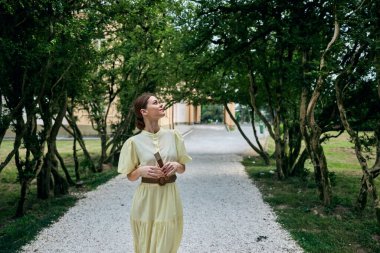 Soluk sarı elbiseli bir kadın yapraklı bir bahçe yolunda yürüyor, parlak bir gün ışığı, yumuşak gölgeler, ve huzurlu bir ruh hali, doğal güzelliği ve rahatlığı ön plana çıkarıyor.