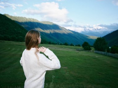 Beyaz örgü süveter giymiş bir kadının olduğu doğa manzarası. Görkemli dağlara ve açık vadiye bakıyor. Seyahat etmeyi, sükuneti, açık havayı ve manzaradan ilham almayı öneriyor..