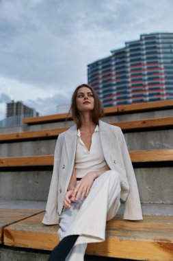 İş kıyafetleri içinde şık bir kadın basamaklarda otururken, arka planda kentsel mimariyle, çağdaş profesyonel bir ruhu somutlaştırarak güven yayar..