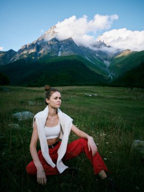Vahşi bir çayırda dinlenen bir kadın, dramatik bir dağ arkası, günlük kıyafetler ve rahat duruş, doğa gösterisi, seyahat ve gün ışığında dinginlik.