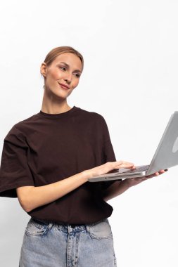 Koyu renk tişörtlü gülümseyen genç bir kadın iki eliyle dizüstü bilgisayar tutuyor, beyaz arka plana yan bakıyor. Teknoloji kavramı, uzaktan çalışma, günlük yaşam tarzı ve dijital iletişim.